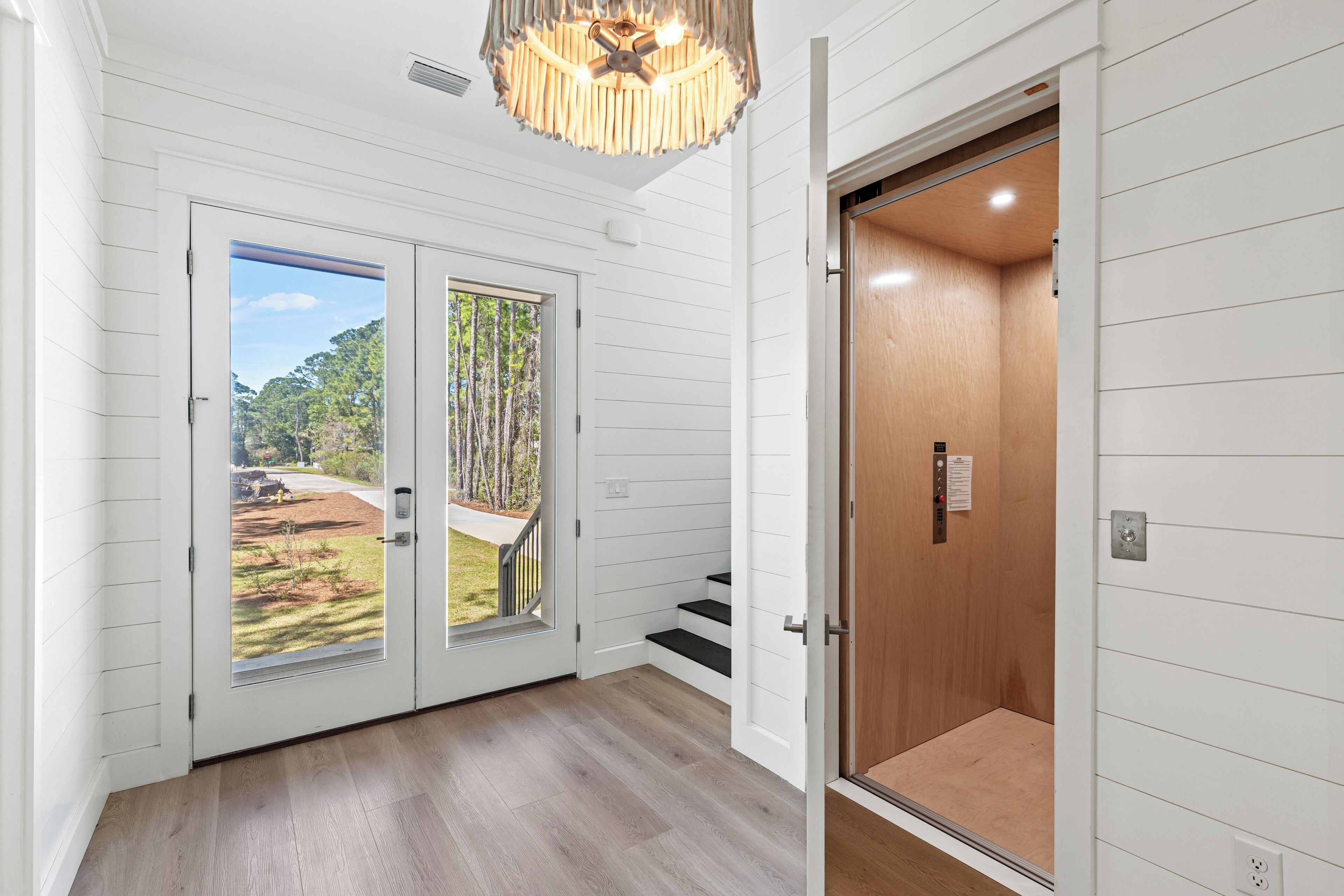 A hallway with a wooden floor, white walls, and a door leading to a balcony with a view of trees and a yard.