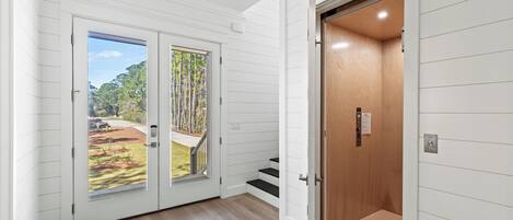 A hallway with a wooden floor, white walls, and a door leading to a balcony with a view of trees and a yard.