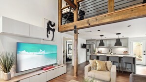 A modern living room with a large flat-screen TV, a sofa, and a dining area with bar stools. - Charming 3BR / 2.5BA Home in  Gentilly – Close to French Quarter & Lakefront (New Orleans)
