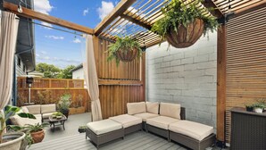A covered outdoor patio with a sectional sofa, hanging plants, and a wooden deck. - Charming 3BR / 2.5BA Home in  Gentilly – Close to French Quarter & Lakefront (New Orleans)