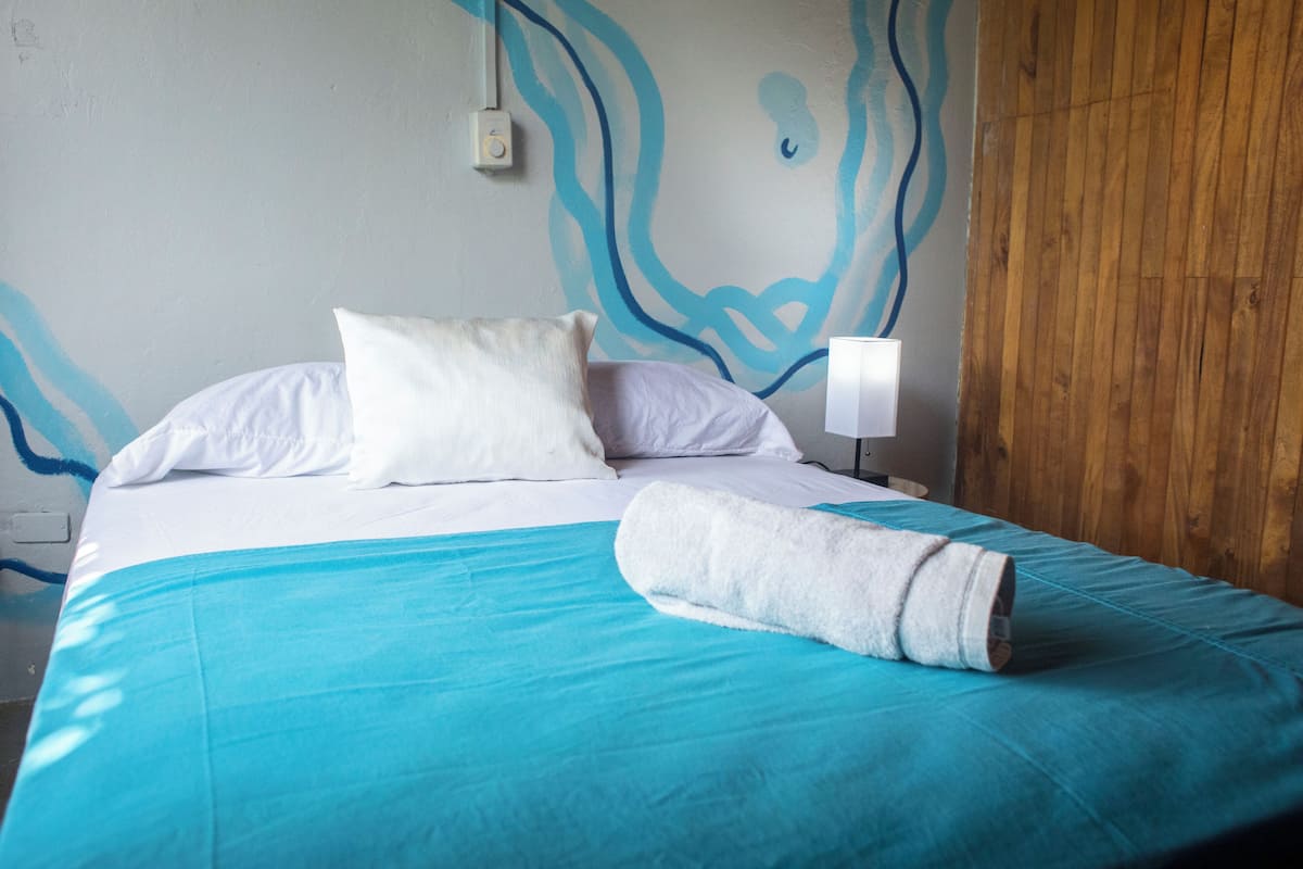 A neatly made bed with a blue bedspread and white pillows, a wooden panel wall, and a simple white bedside lamp.