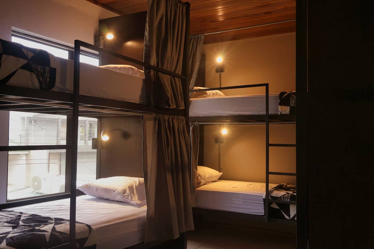 A dormitory room with bunk beds, a window, and a nightstand with a lamp.