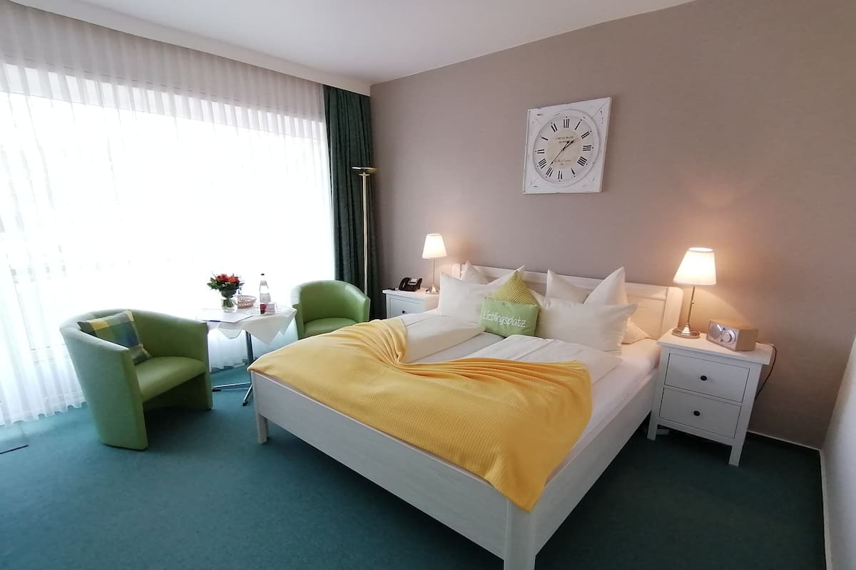 Une chambre d’hôtel avec un lit, deux tables de chevet, une chaise, une table ornée d’un vase de fleurs et une horloge au mur.