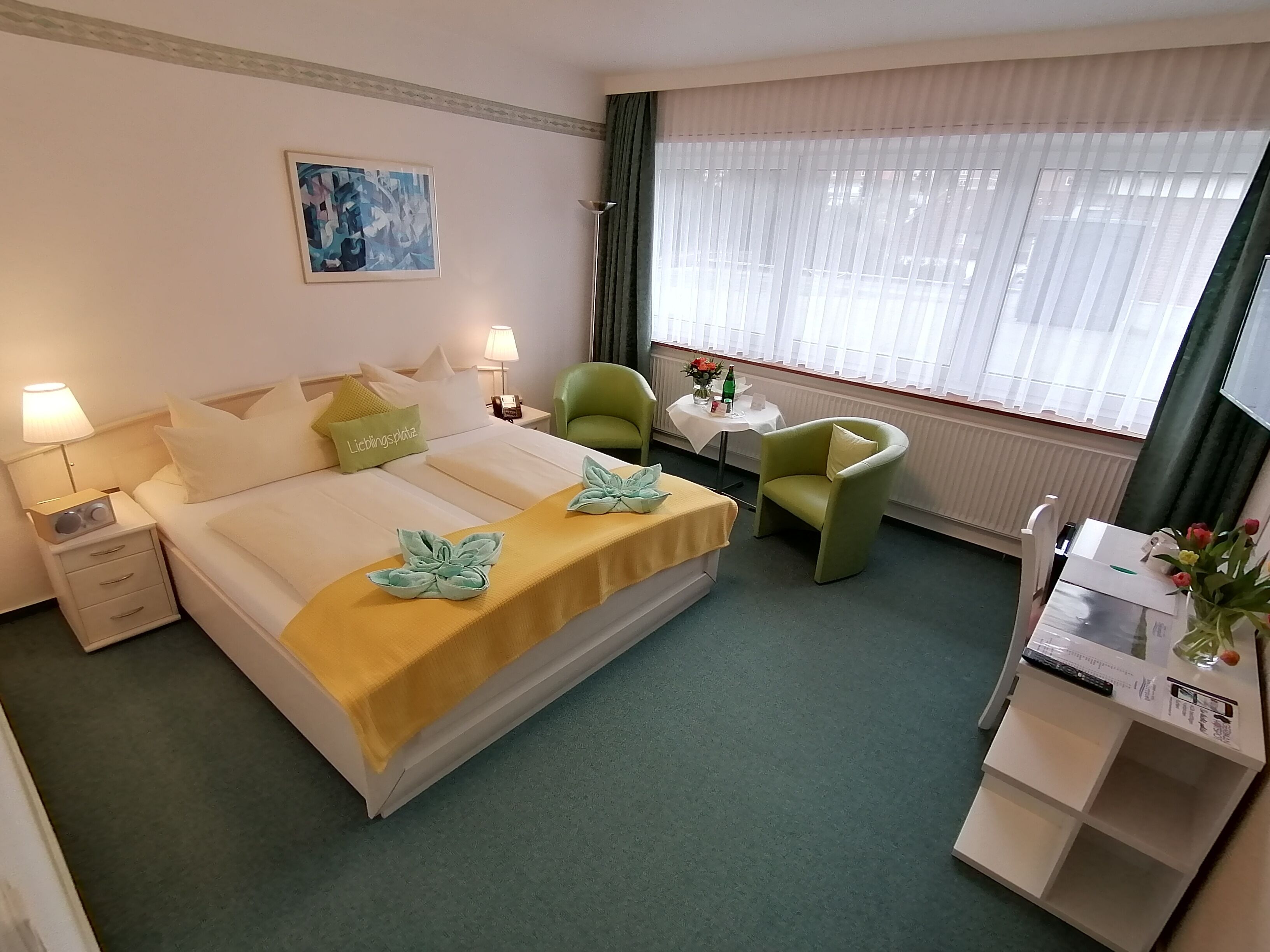 Une chambre d’hôtel avec un lit, deux chaises, un bureau, une lampe, un tableau et une fenêtre avec des rideaux.