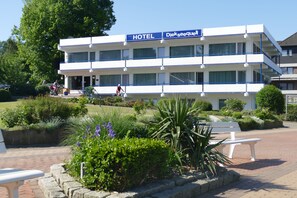 A hotel with a sign reading "HOTEL Diekseequell". It features a multi-story building with balconies, surrounded by well-maintained gardens and a paved walkway.