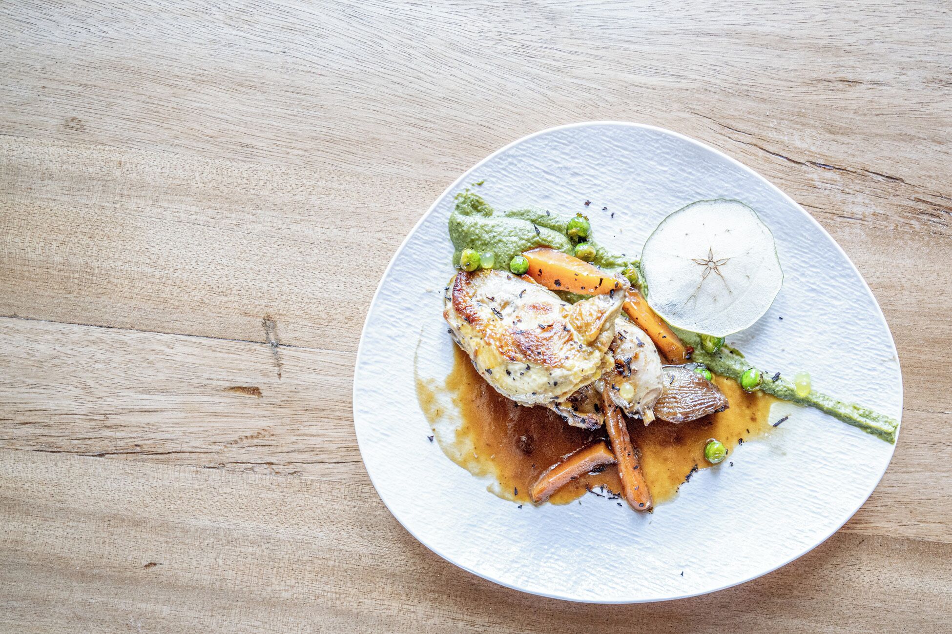 A plate with a roasted chicken, vegetables, and a slice of apple on a textured white plate.
