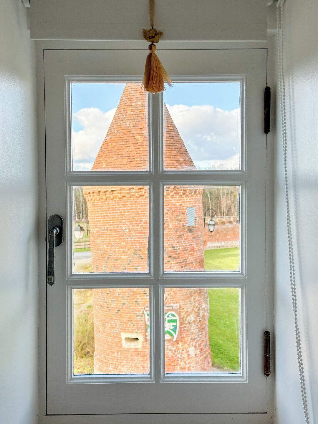 Eine weiße Tür mit einem Fenster, das in vier Felder unterteilt ist, zeigt ein Backsteingebäude mit einem Kegeldach und einem grünen Rasen davor.