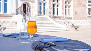 Une table dressée à l’extérieur, avec un verre de jus d’orange, une assiette et des couverts, devant un bâtiment historique doté d’un magnifique escalier.