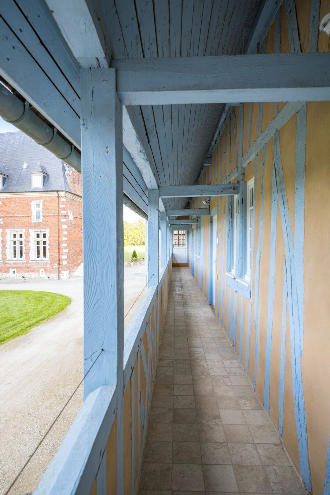 Un couloir couvert doté d’un sol carrelé, de poutres en bois et offrant une vue sur un bâtiment en briques avec des fenêtres.
