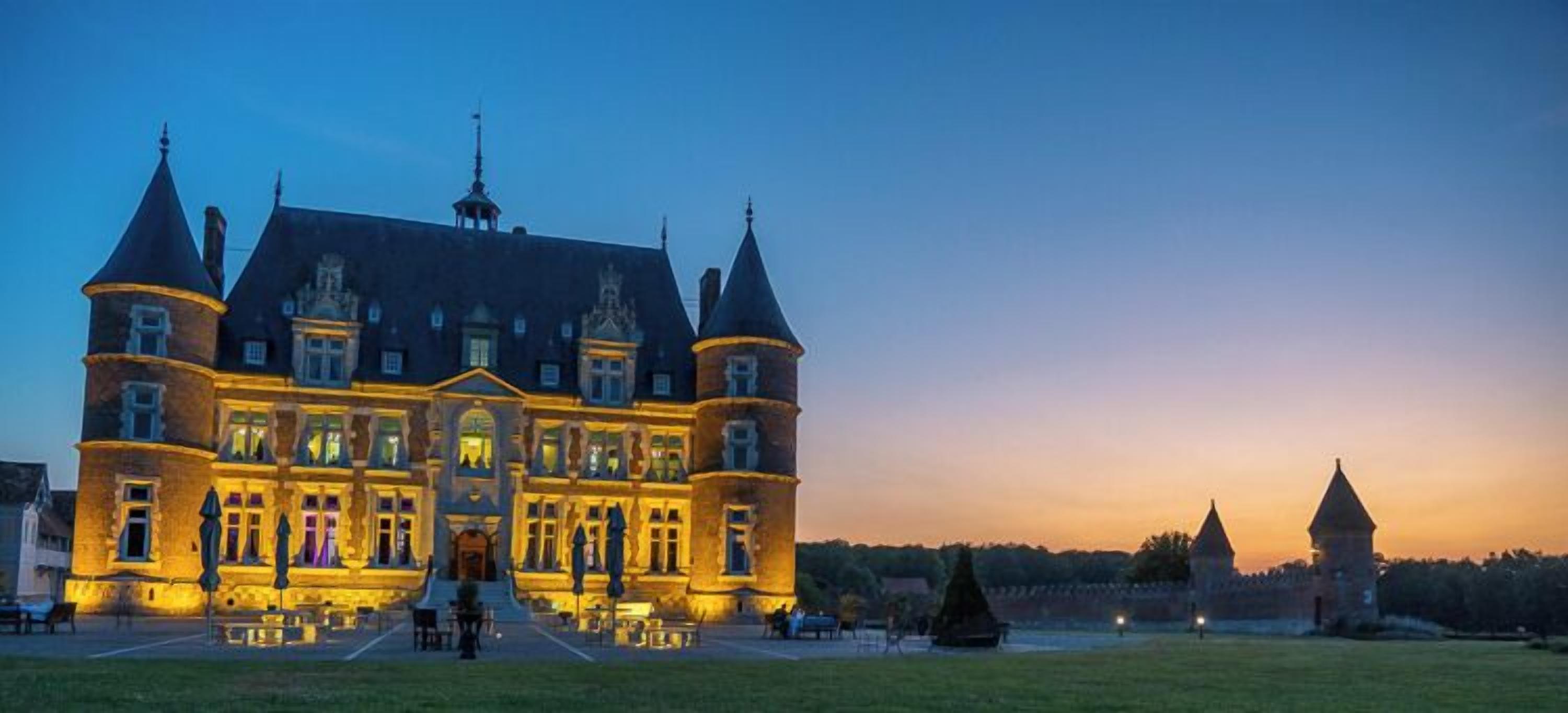 A large, historic building with multiple towers and a symmetrical facade, featuring a central entrance and a well-maintained lawn in front.