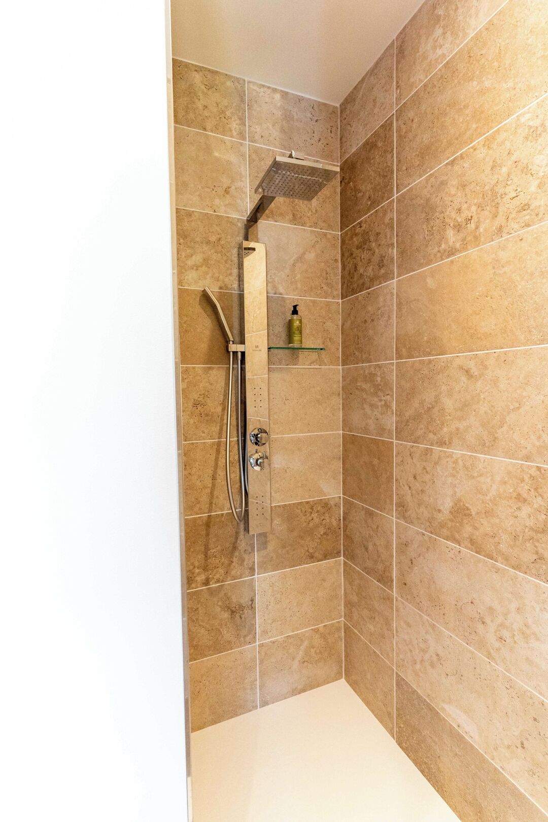 Un espace douche équipé d’un pommeau de douche à main, d’une étagère en verre sur laquelle est posée une bouteille, et de murs recouverts de carreaux.