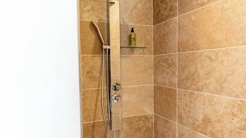 Un espace douche équipé d’un pommeau de douche à main, d’une étagère en verre sur laquelle est posée une bouteille, et de murs recouverts de carreaux.