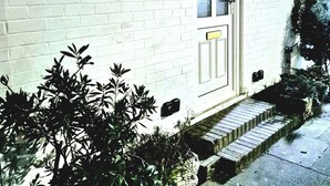 Ein weißes Backsteingebäude mit einer Haustür und Stufen, die zum Eingang führen, sowie ein kleiner Gartenbereich mit Sträuchern.