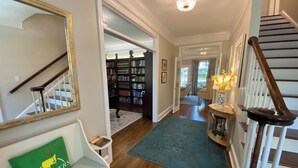 A spacious hallway with a wooden staircase, a green couch with a 'Masters' pillow, a bookshelf, and a chandelier. - Masters Tournament Rental; 4 Bed, 2.5 Bath-4 Bedroom Home in Heart of Evans, GA (Evans)