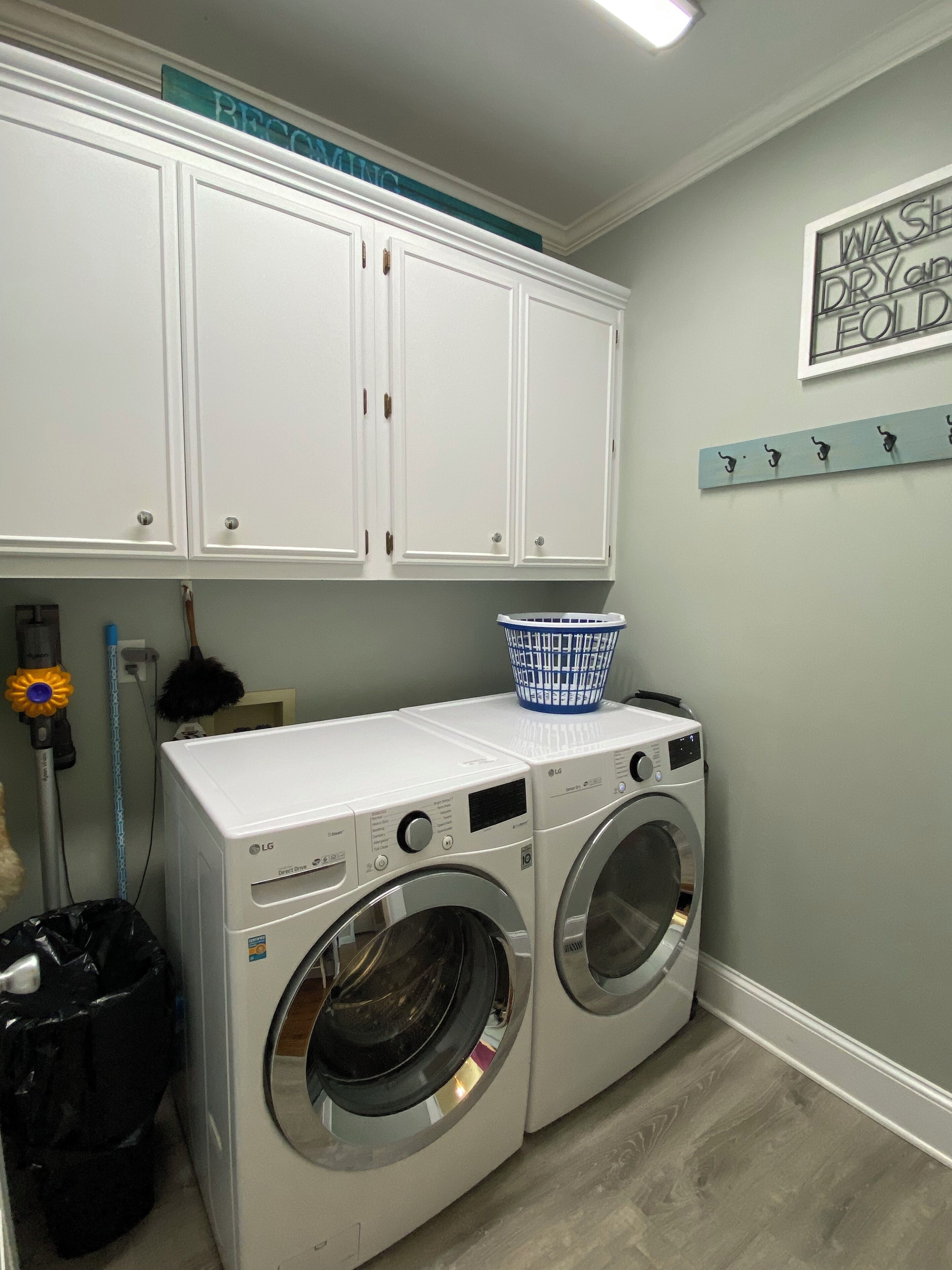 A laundry room with two front-loading washing machines, a blue laundry basket, a black trash bag, a vacuum cleaner, and a wall-mounted drying rack.
