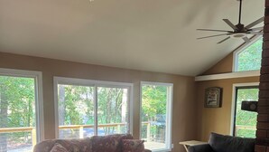 Ein geräumiges Wohnzimmer mit dunklem Holzboden, einem blumigen Sofa, einem dunkelbraunen Ledersessel und einem Holztisch. Große Fenster bieten einen Blick ins Freie.