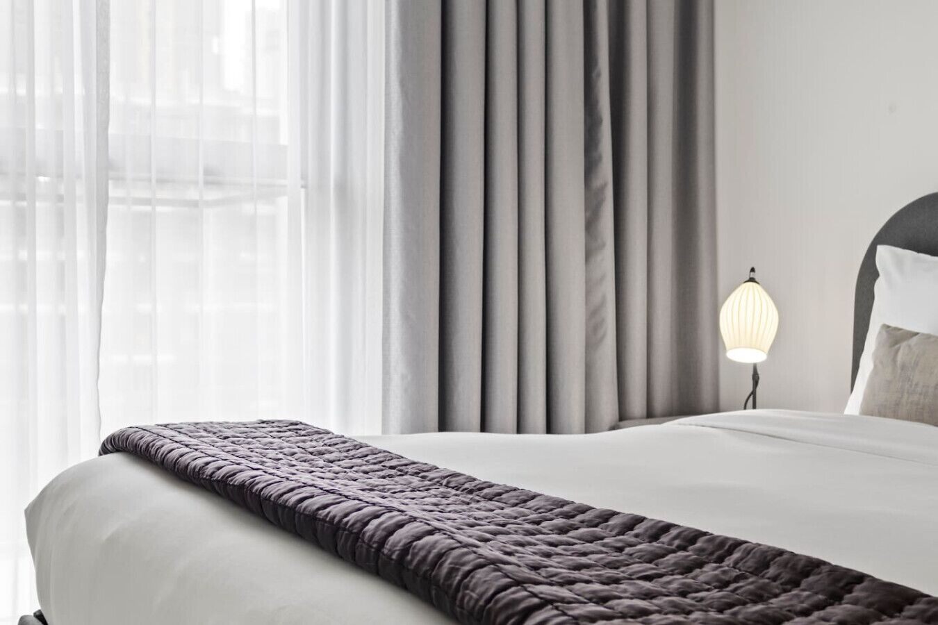 Een net opgemaakt bed met een donge deken, een bedlampje en een raam met dunne gordijnen.