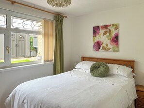 A bedroom with a bed, a window with curtains, a wooden headboard, and a wall-mounted painting. - A charming bungalow in Port Eynon, Gower, just minutes from the beach. (Port Eynon, Gower Peninsula)