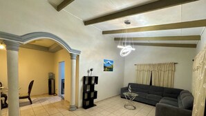 Un salón con sofá gris, una mesita de centro pequeña y una escultura de vidrio decorativa. El piso es de baldosas y el techo tiene vigas a la vista.