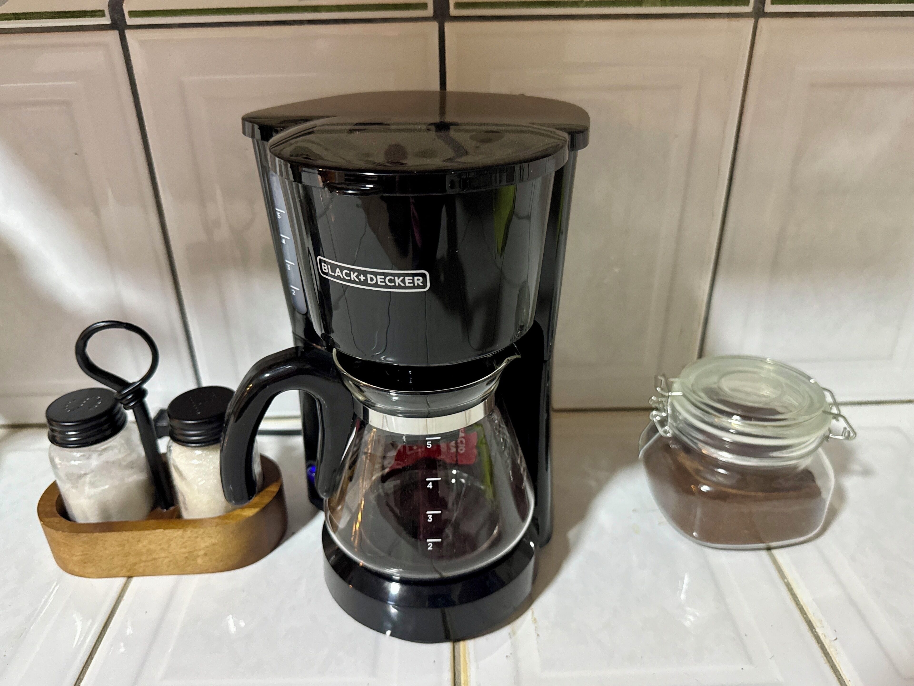 Una cafetera negra con jarra de vidrio, un estante de madera para especias con frascos, y un tarro de azúcar en una encimera de cocina con baldosas.