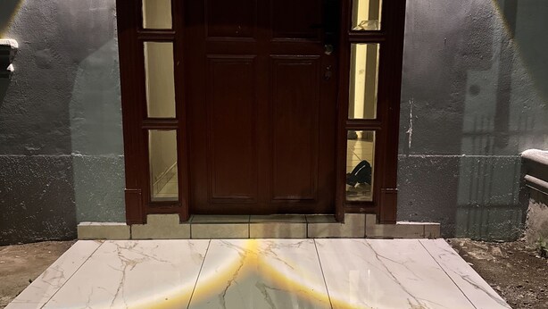 Una puerta de madera con paneles de vidrio, flanqueada por dos luces fijadas a la pared, que da acceso a un área de entrada con baldosas.