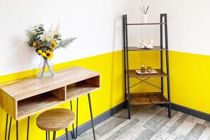 Ein Eckzimmer mit Holztisch, kleinem runden Tisch und einem schwarzen Metallregal an einer gelben Wand.