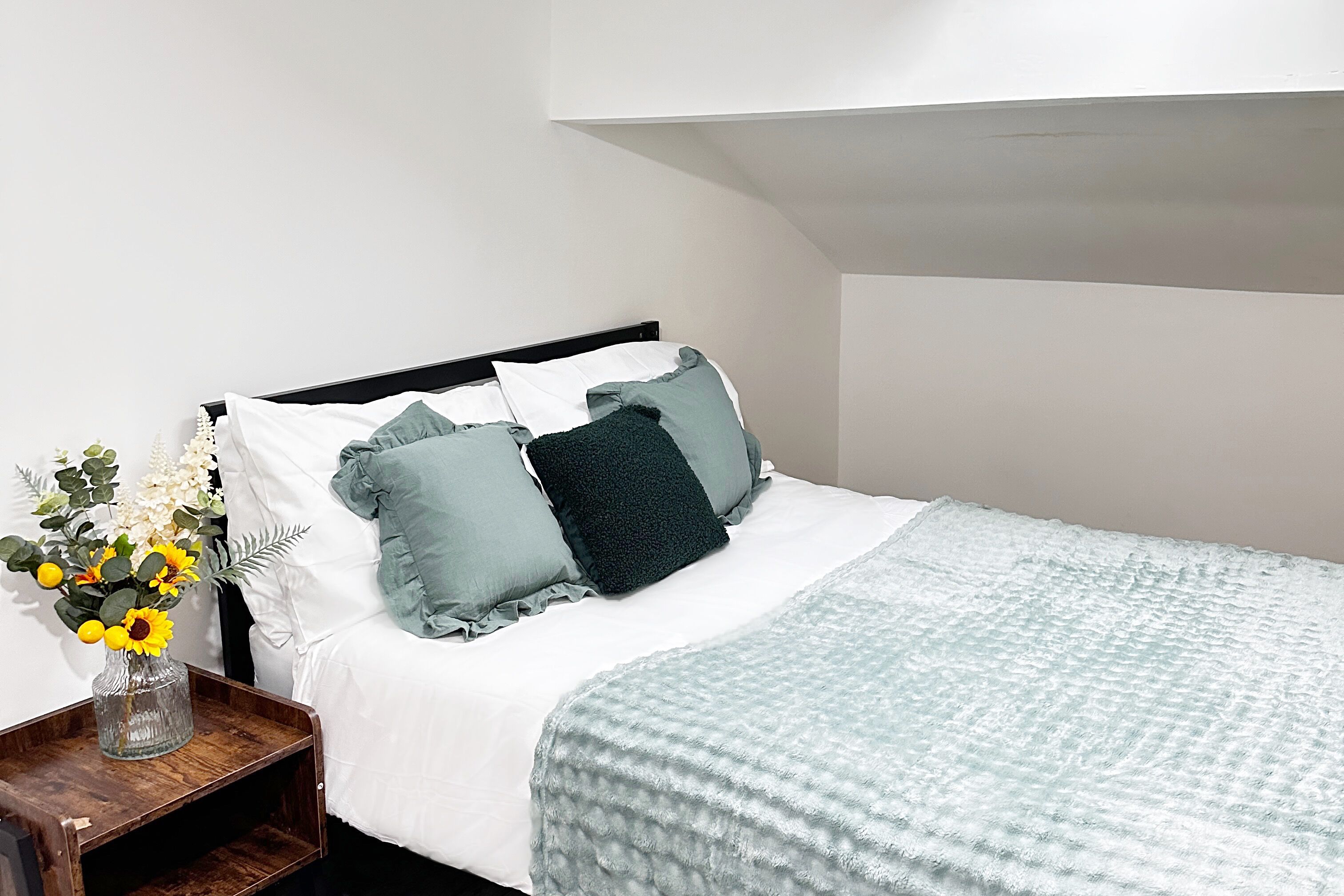 Un lit bien fait, recouvert d’une couette blanche et turquoise, avec deux oreillers et un vase de fleurs posé sur une table de chevet en bois.