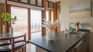 A modern kitchen with a large window, a round dining table, and a vase of dried flowers. - Luxe Oceanfront w/ HotTub | Icon of Mission Beach (San Diego)