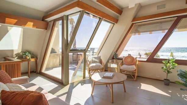 A sunlit room with a large window overlooking the sea, furnished with a sofa, chairs, and a small table. - Luxe Oceanfront w/ HotTub | Icon of Mission Beach (San Diego)