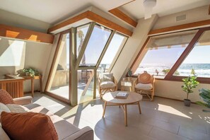 Une pièce lumineuse, baignée de soleil, avec une grande fenêtre donnant sur la mer, et meublée d’un canapé, de fauteuils et d’une petite table.