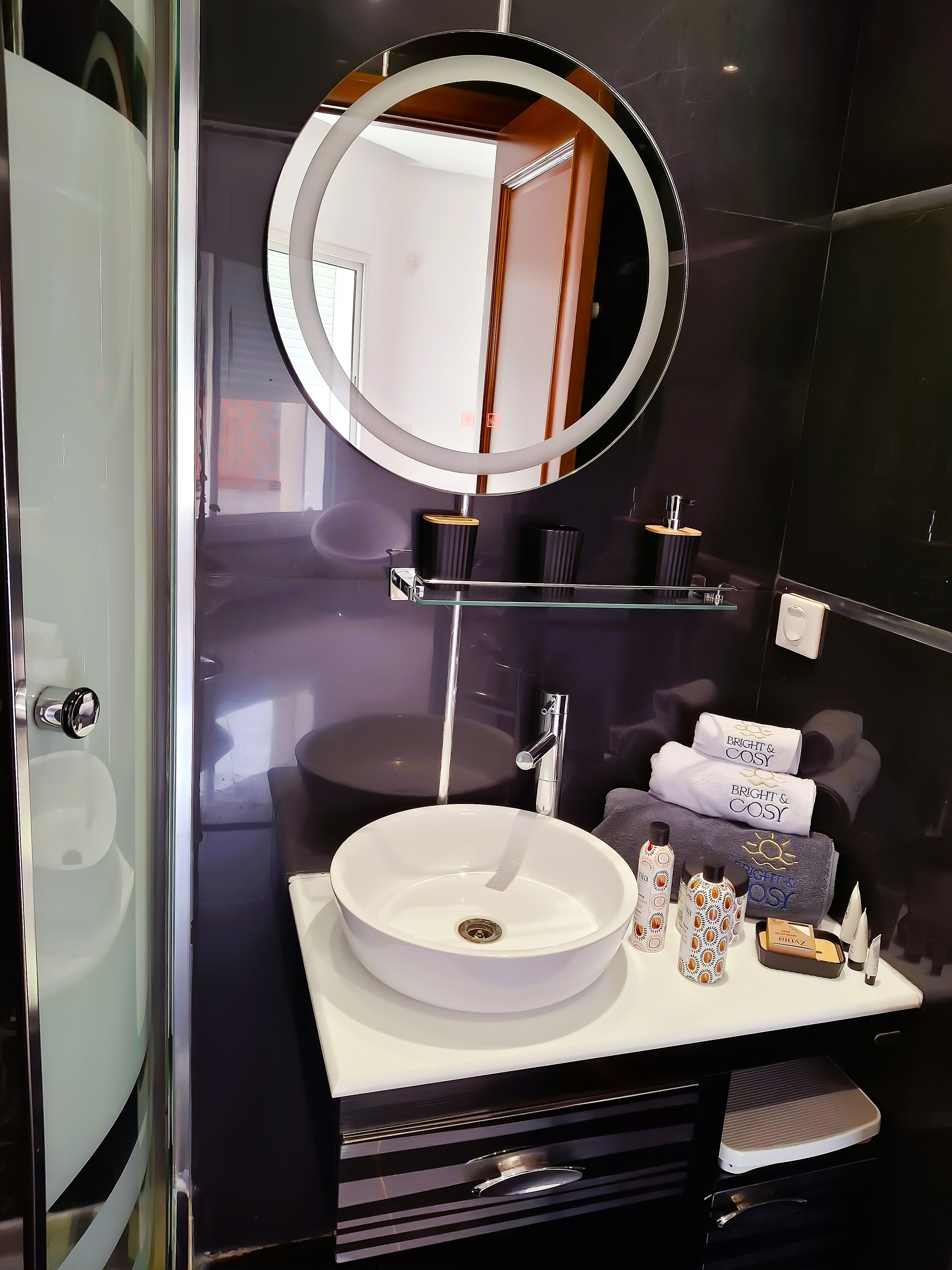 Une salle de bain moderne avec un miroir rond, un lavabo blanc et des articles de toilette posés sur le meuble-lavabo.