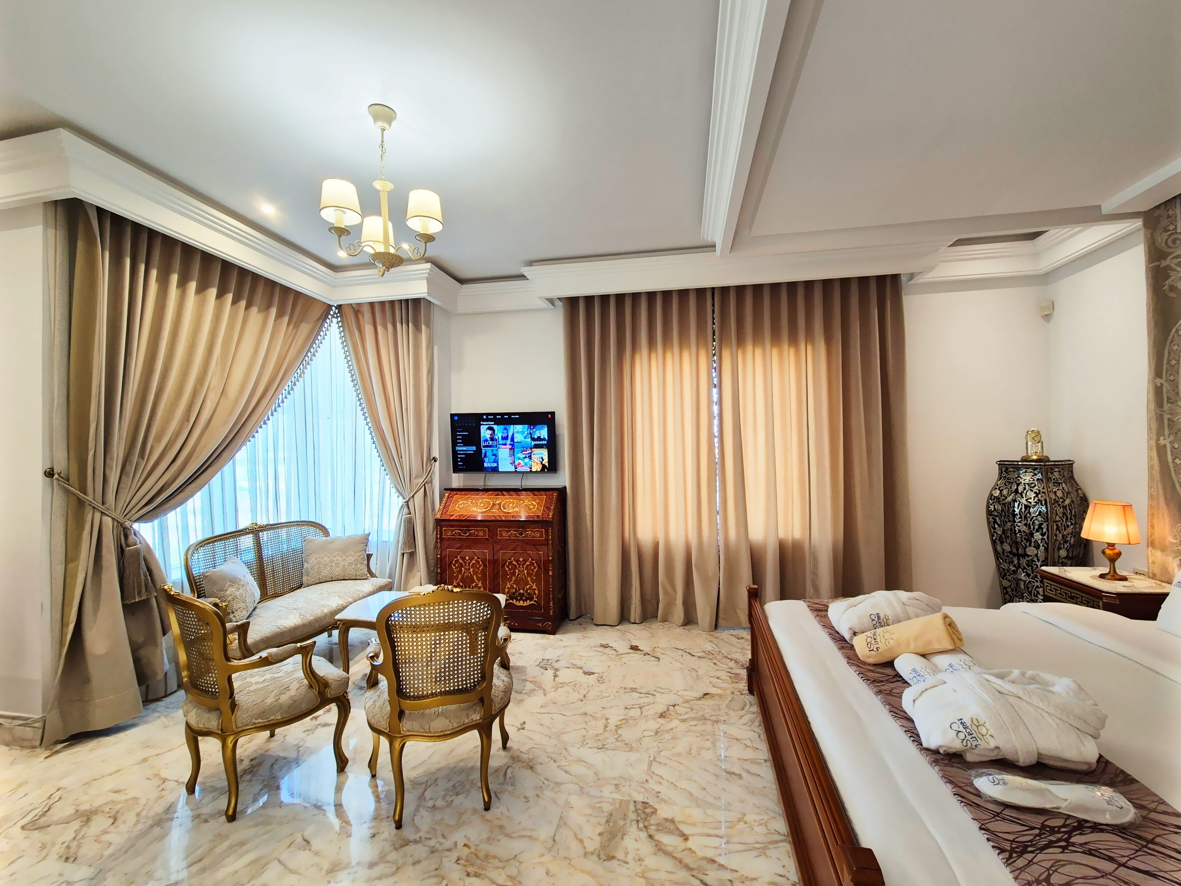 Una habitación de hotel con suelo de mármol, cama con ropa blanca, sofá, mesa de comedor con sillas, televisión y un jarrón decorativo.