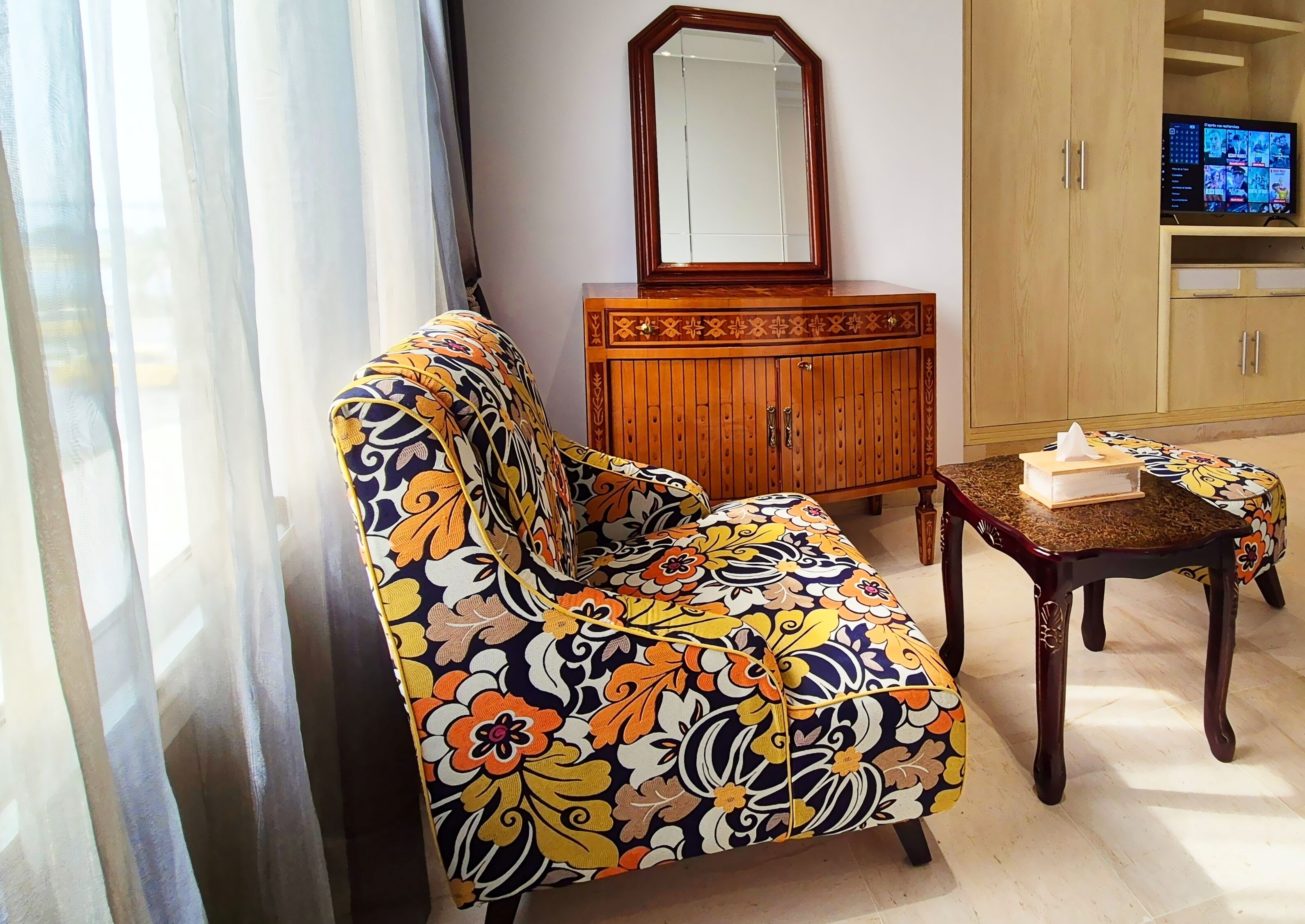 Una sala de estar con un sofá floral, un mueble de madera, una mesita con una caja de pañuelos y un televisor sobre un soporte de madera.