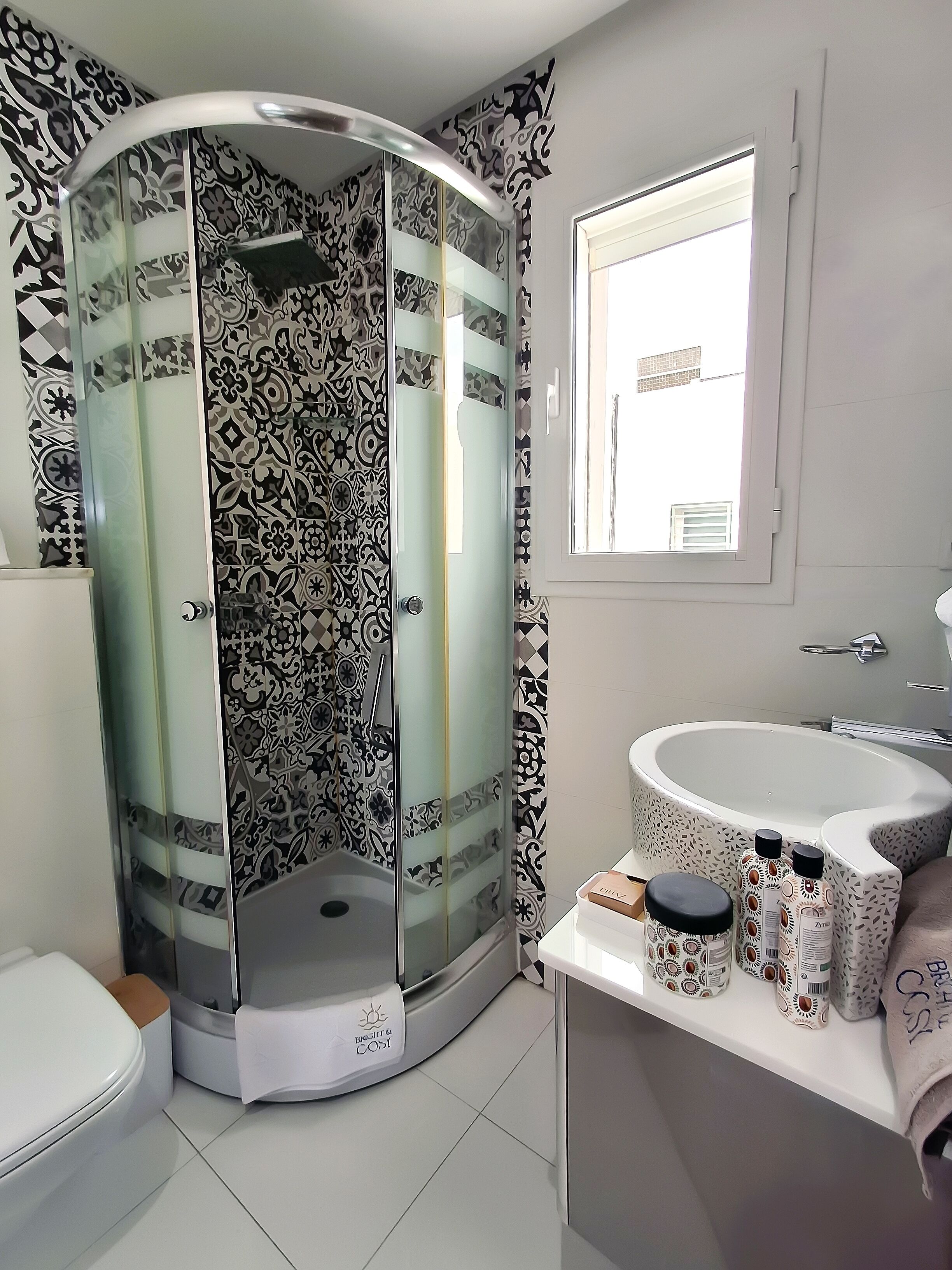 Un baño con ducha envuelta en vidrio, azulejos con diseño, un lavamanos con botellas decorativas y una ventana.