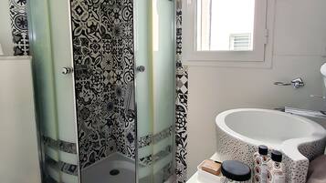 Un baño con ducha envuelta en vidrio, azulejos con diseño, un lavamanos con botellas decorativas y una ventana.