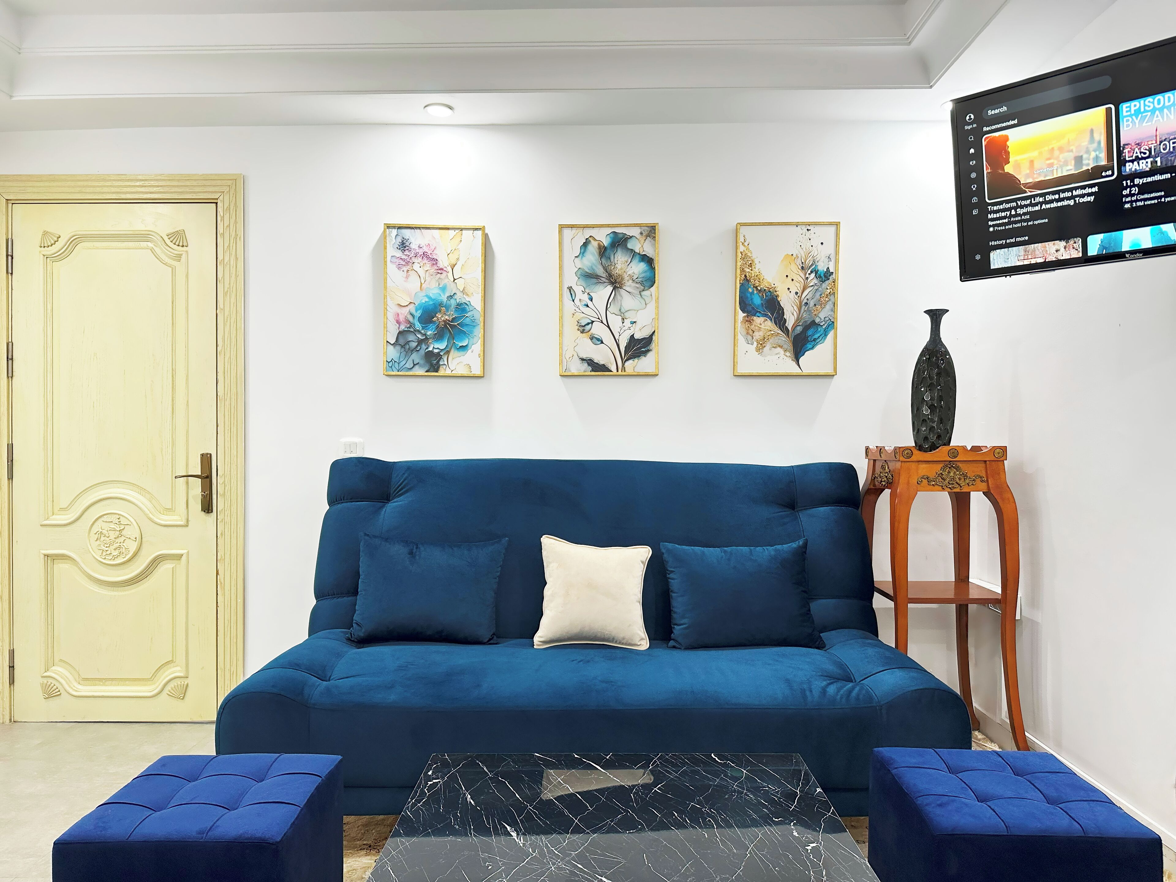 Een woonkamer met een blauwe bank, twee blauwe poufs, een zwarte salontafel, en drie abstracte bloemschilderijen aan de muur.