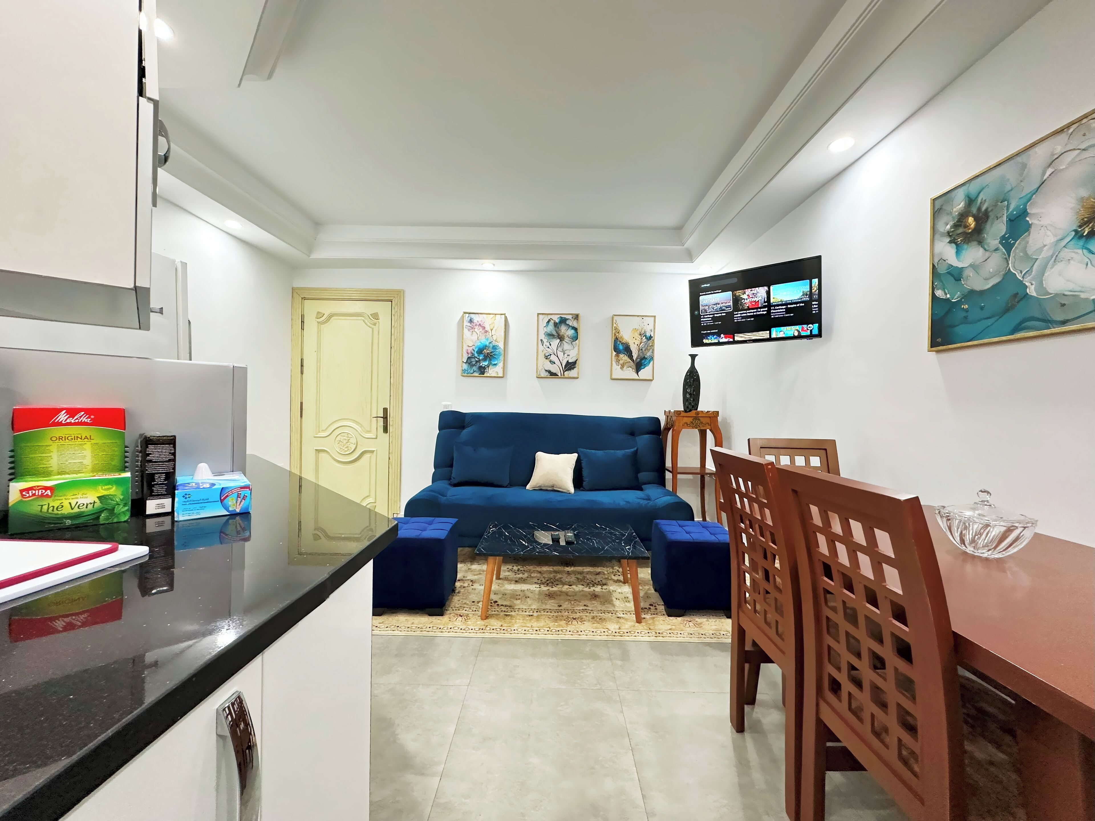Un espace de vie compact comprenant un coin cuisine, un canapé bleu, une table à manger et un téléviseur fixé au mur.