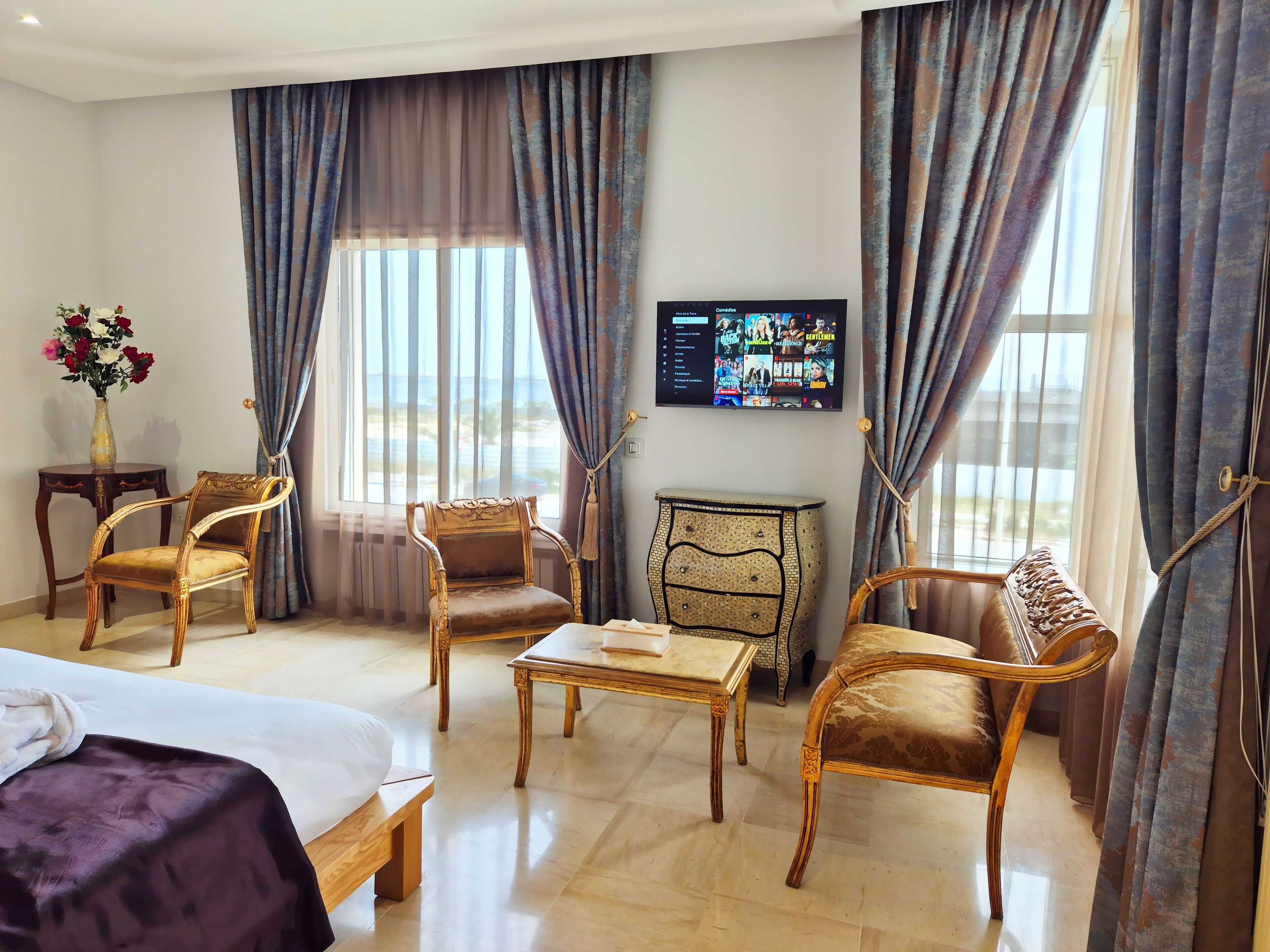 Quarto de hotel com cama, duas poltronas, uma mesa pequena, mesa lateral com um vaso de flores e televisão na parede.