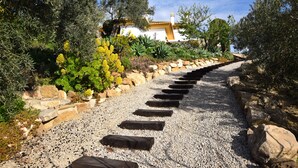 Un chemin mène à un bâtiment à la façade blanche et au toit jaune, entouré de différentes plantes et d’arbres.