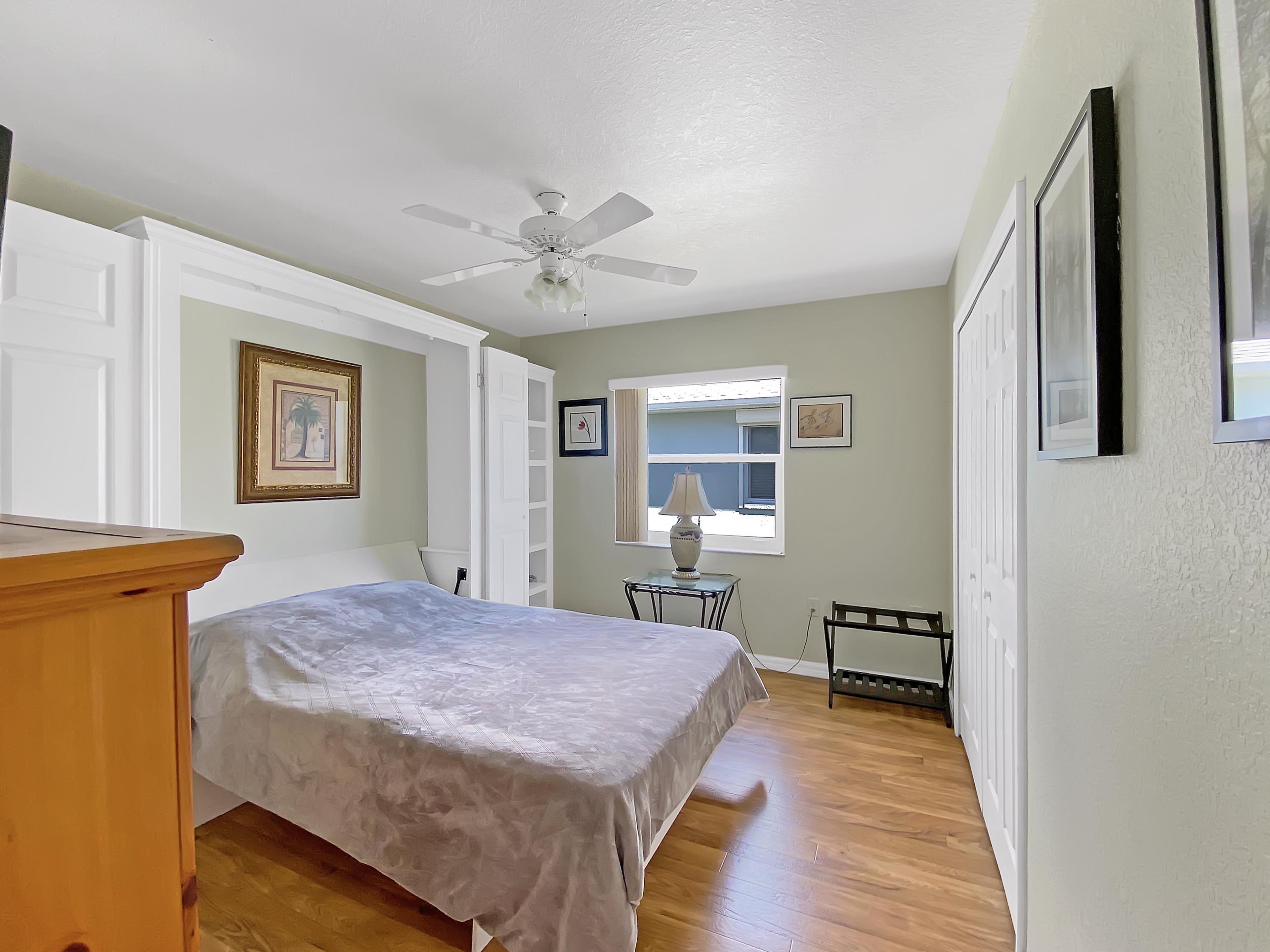 A bedroom with a bed, a ceiling fan, a window with a view, a table with a lamp, and a chair.