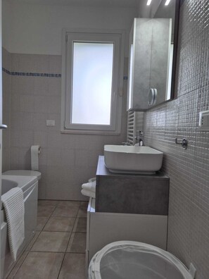 Ein modernes Badezimmer mit einer Badewanne, einem Waschbecken und einer Toilette. Ein Fenster sorgt für natürliches Licht, und über dem Waschbecken befindet sich ein Spiegel.
