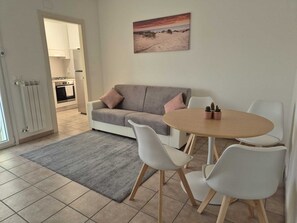 Ein modernes Wohnzimmer mit einem grauen Sofa, weißen Stühlen, einem runden Holztisch und einem Gemälde mit Strandmotiv.