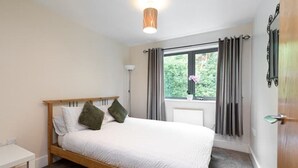 Un dormitorio con cama de madera, ropa de cama blanca, una mesita de noche, una lámpara de pie, una ventana con cortinas y un radiador.