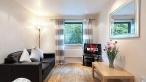 Una sala de estar con un sofá negro, una mesa de centro de madera, un televisor mostrando el logotipo de Netflix y una ventana con cortinas.