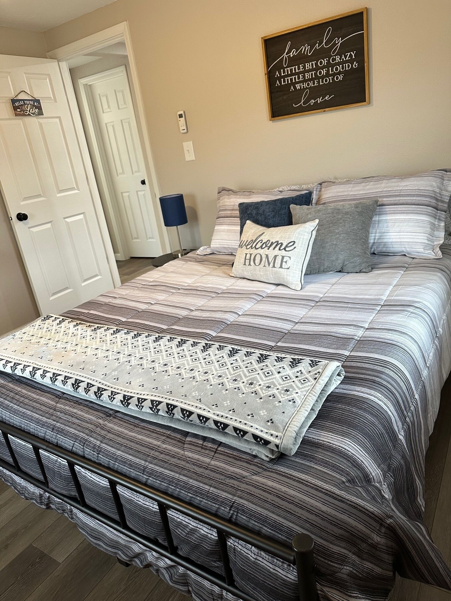 A neatly made bed with striped bedding and a welcome home pillow, a framed family quote on the wall, and a door to the left.