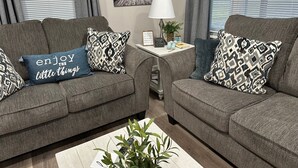 A living room with two grey sofas, patterned throw pillows, a small table with a lamp, a framed picture on the wall, and a plant on the floor. - Cozy 2-bedroom cabin with WiFi, AC in tranquil Moravia (Moravia)