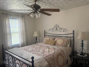 Quarto com cama, ventilador de teto, janela com cortinas e mesa de cabeceira com abajur.