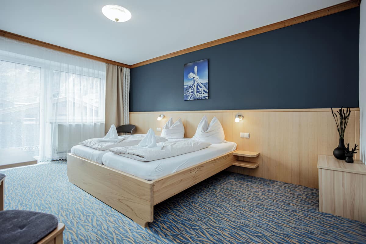Una habitación de hotel con una cama de madera, un ventanal con cortinas, un cuadro en la pared y una alfombra azul estampada.