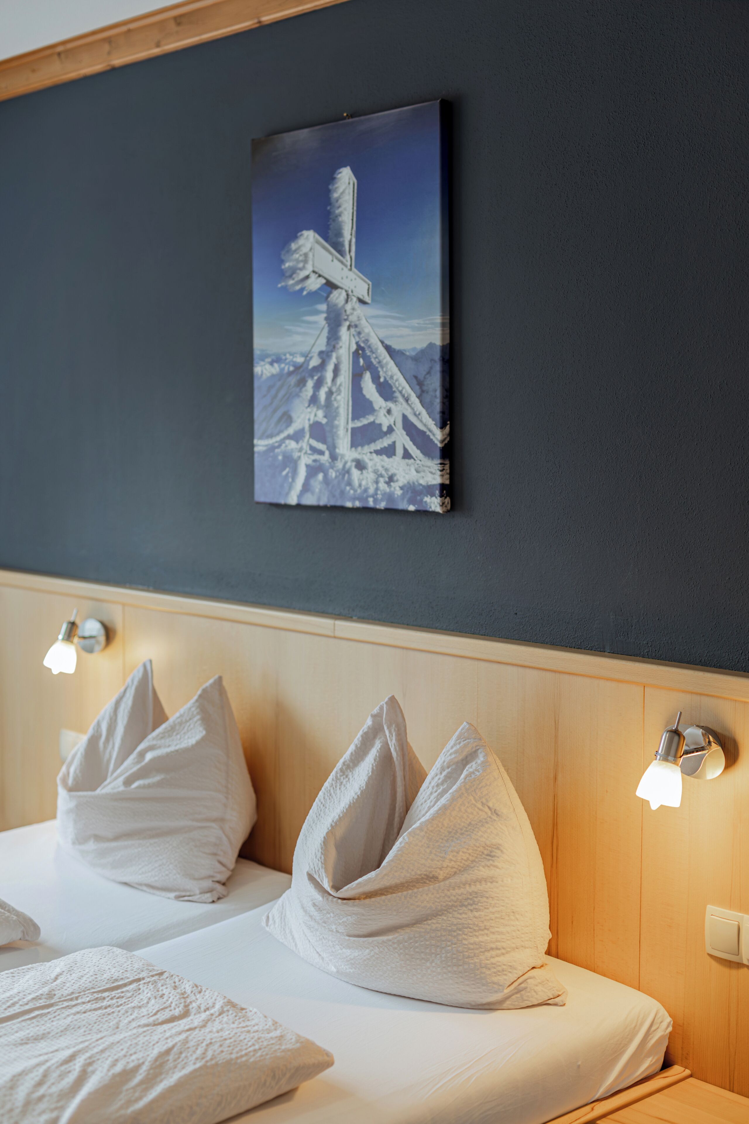 A hotel room with a bed, white pillows, a painting of a snowy landscape, and wall-mounted lights.