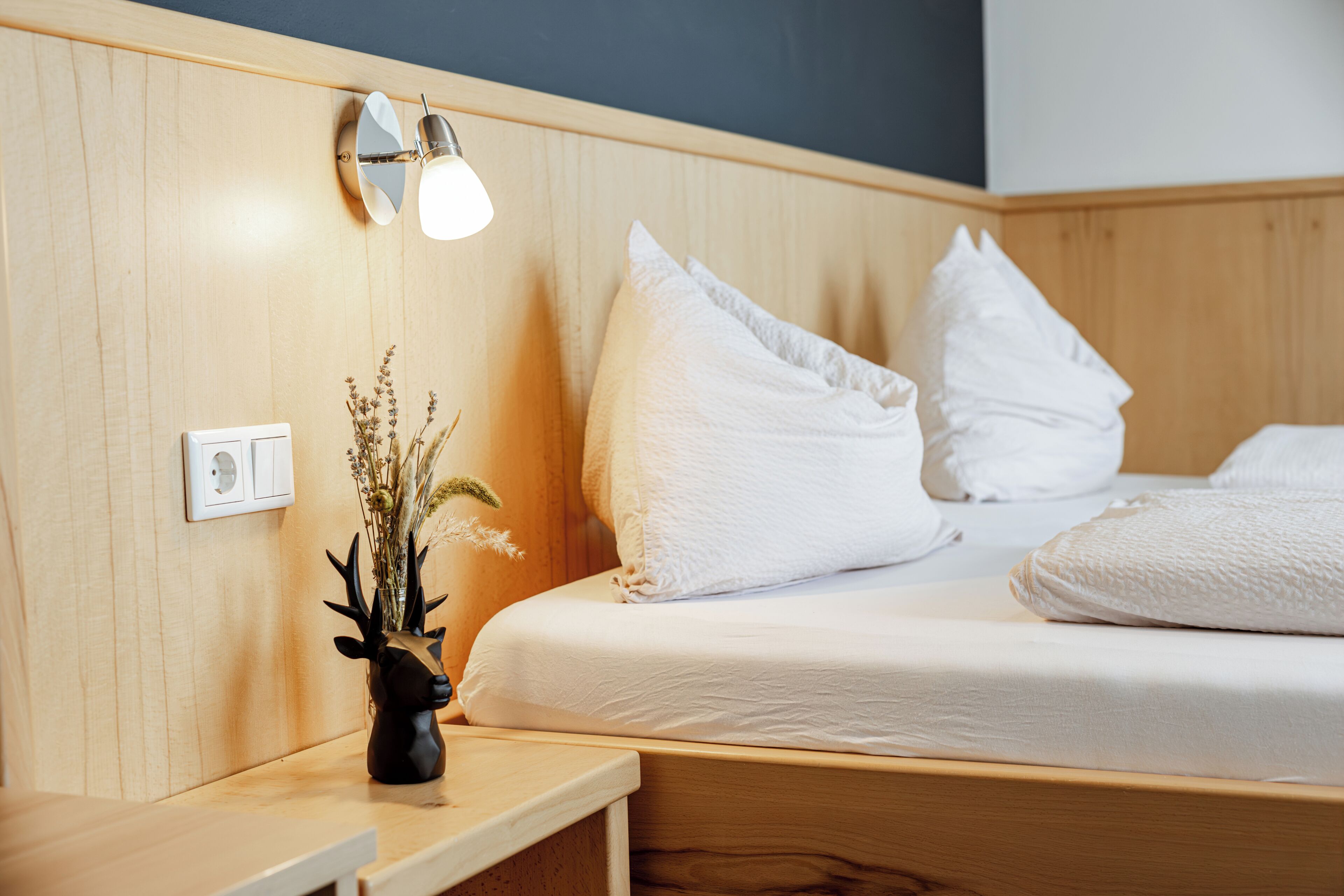 A neatly made bed with white linens and pillows, a bedside table with a lamp, and a wall-mounted light fixture.